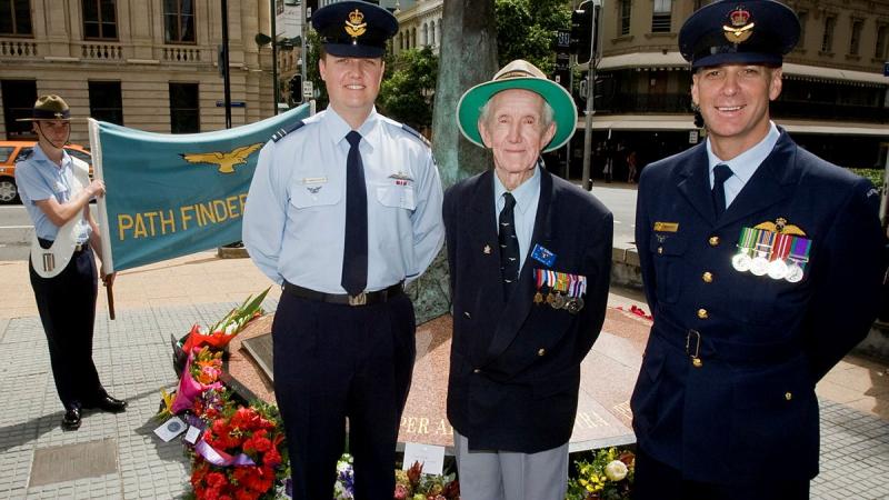 Two military members stand with a decorated veteran in front of a monument and a person holding a banner.