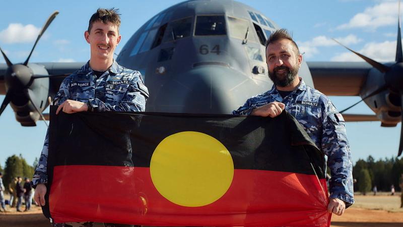 Two military men hold a First Nations flag in front of an aircraft.