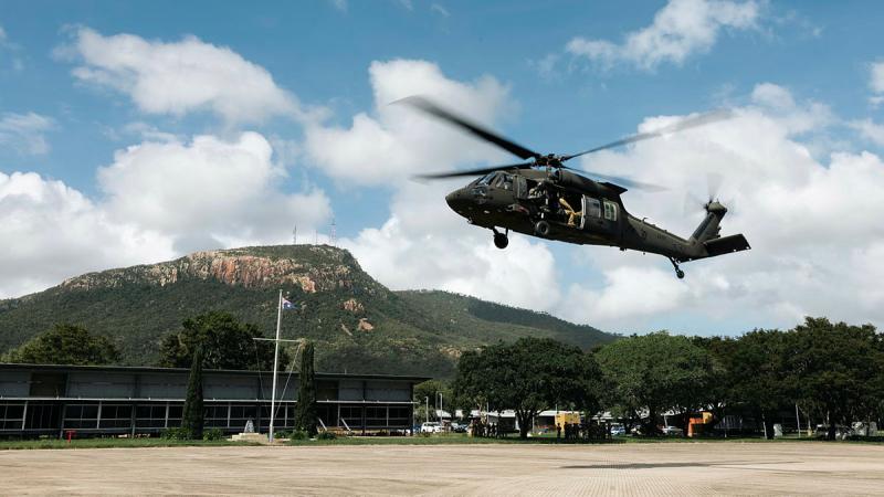 Helicopter flies on partly cloudy day, with hill in background.