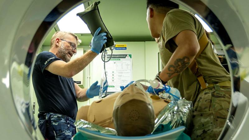 Defence medical personnel are seen through the tunnel of a scanner. One holds a fluid bag and another tends to a 'dummy' patient.