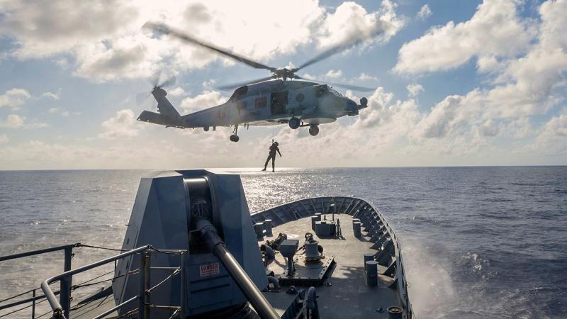 A person hangs from a helicopter over the end of a ship while on the ocean.