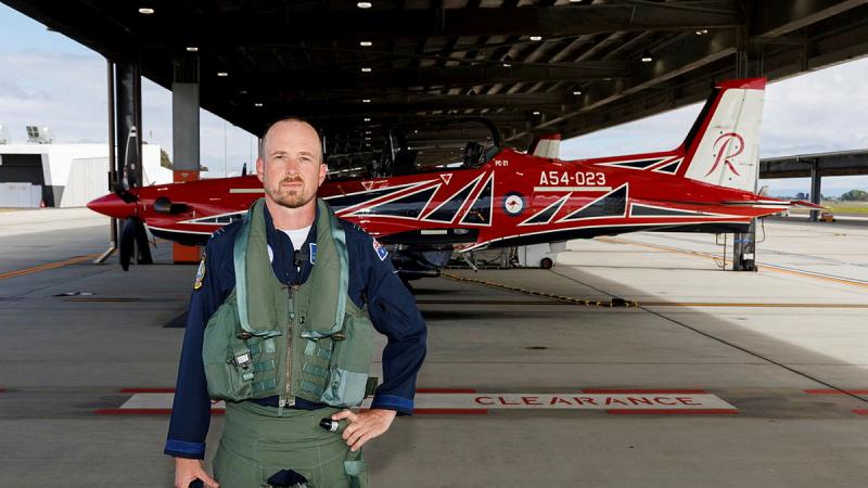 An Air Force pilot standing undercover on the tarmac in front of a red aircraft.