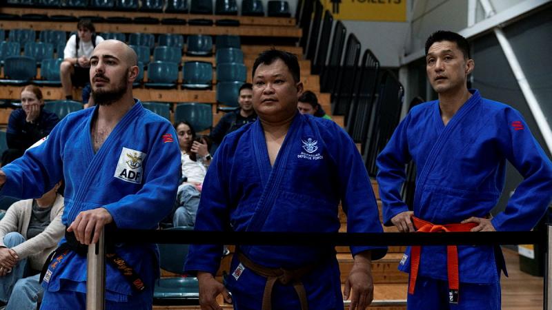 Three men in judo uniforms standing side by side watching a match. 