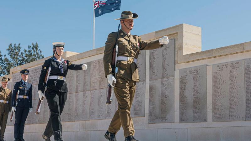 Personnel march in formation past a memorial site and the Australian flag.