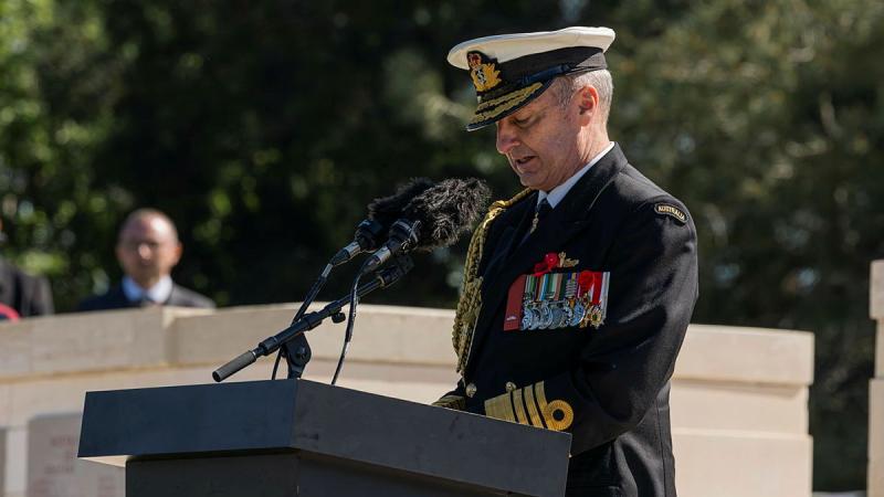 A man in formal Navy uniform speaks at a podium.