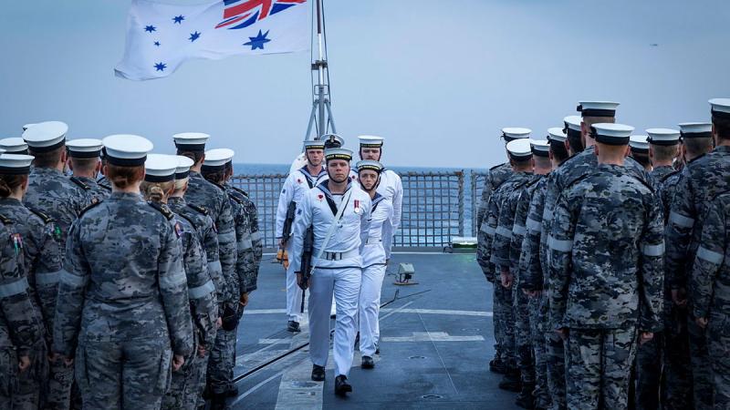 Four sailors march between two blocks of sailors on parade with a white ensign in the background