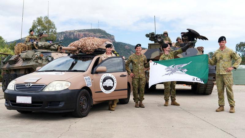 Military personnel stand with three modified vehicles, a flag and a large black bird.