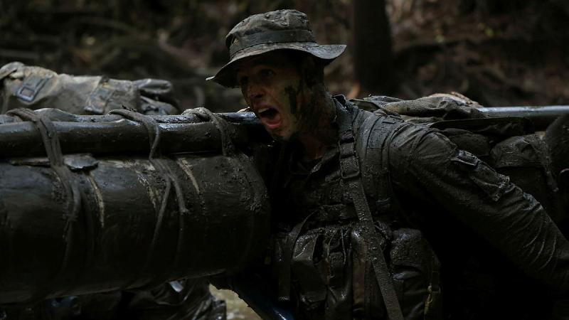 A soldier in the jungle helping to carry a large litter on his shoulder.