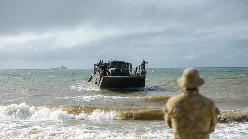 A solider stands on the beach as a landing craft approaches from the sea.