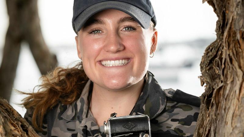 A female sailor leans into a tree trunk and smiles while holding a camera.