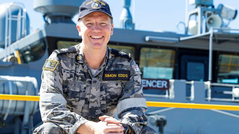 A Navy member sits smiling with hands clasped near a railing, in front of a ship.