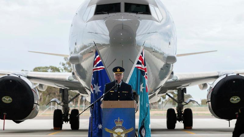 A man in military uniform speaks at a podium, with a large aircraft in the background.