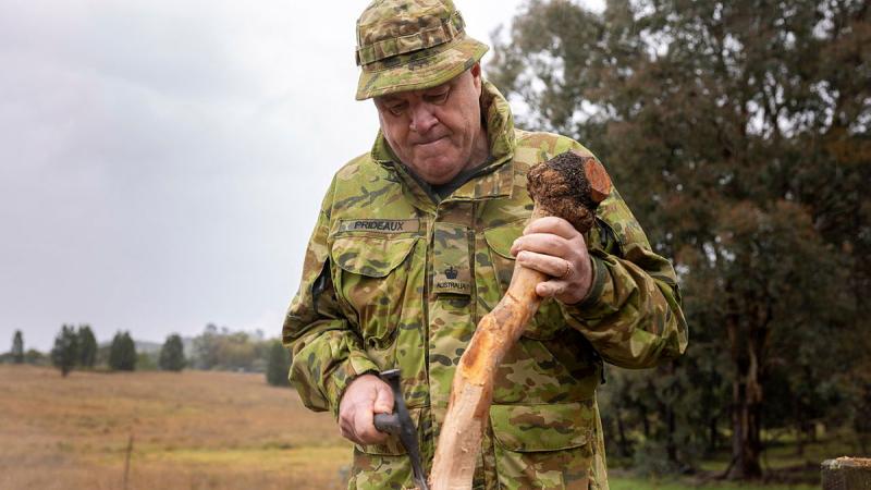 A man in camouflage uniform uses a small axe to shape a piece of wood.