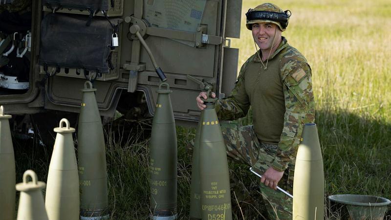 A soldier kneels and holds a piece of ASP Huntsman ammunition near the back of an Army vehicle.