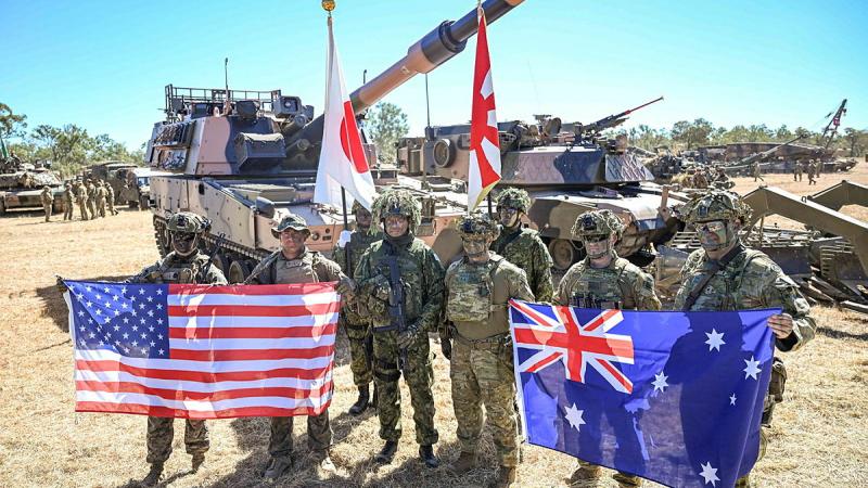 Military personnel hold up their countries flags while standing in front of military vehicles.