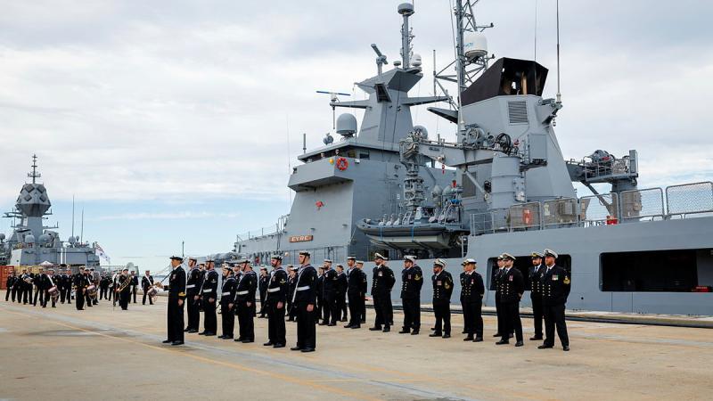Sailors stand in formation on a concrete surface in front of a docked ship.