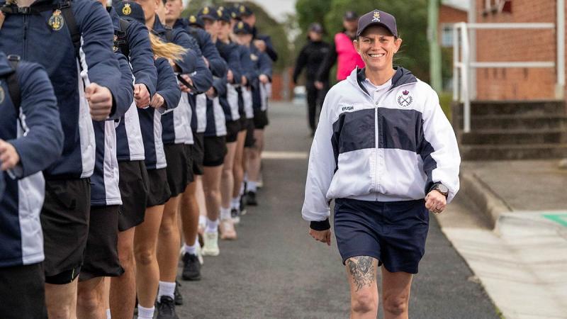 A woman in Navy exercise gear marches alongside a line of recruits in exercise uniform.