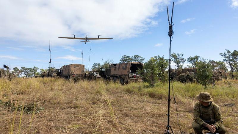 A small aircraft flies from behind towards a man in Army uniform crouching in front of a laptop computer.