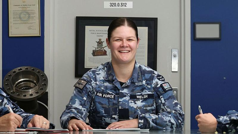 A military woman sits at a table with two others (unseen) taking notes.