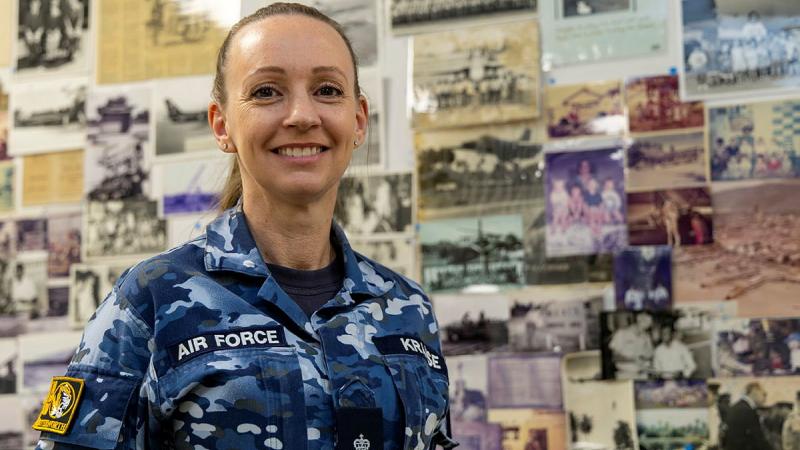 A female Air Force member stands, smiling, in front of a  display board of old photos.