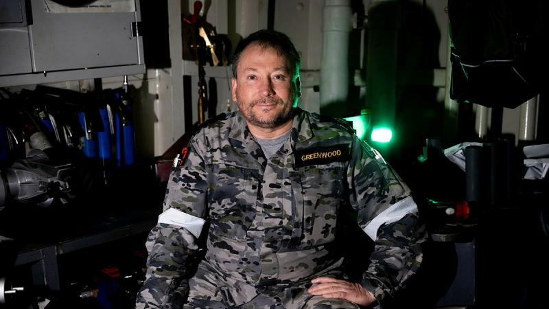 A Navy member in uniform sits with one hand on his hip in a room of equipment.