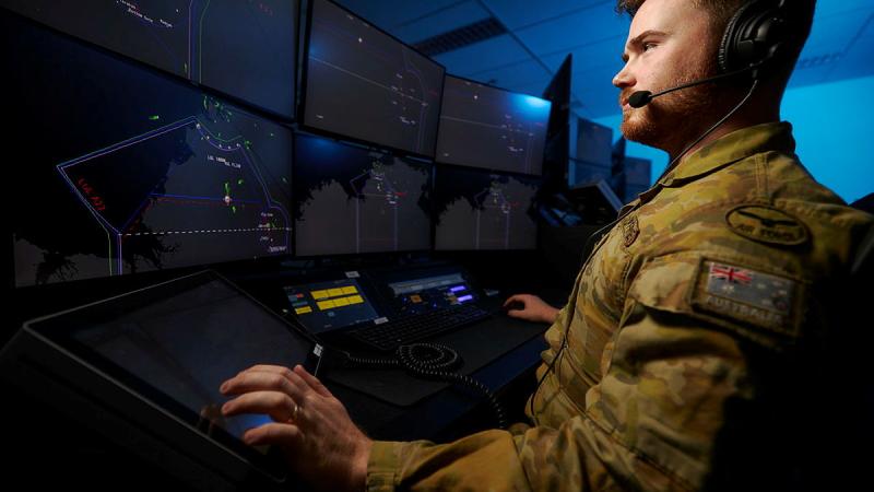 A uniformed man sits at a workstation with multiple computer screens.