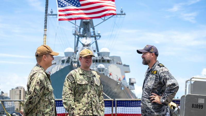 Three sailors speak to each other in front of a flag.