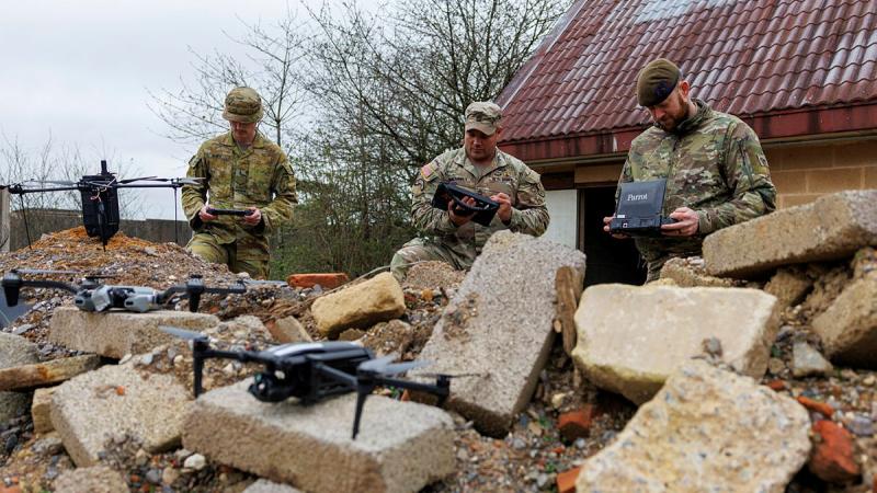 Army personnel standing in concrete rubble hold controls for drones.