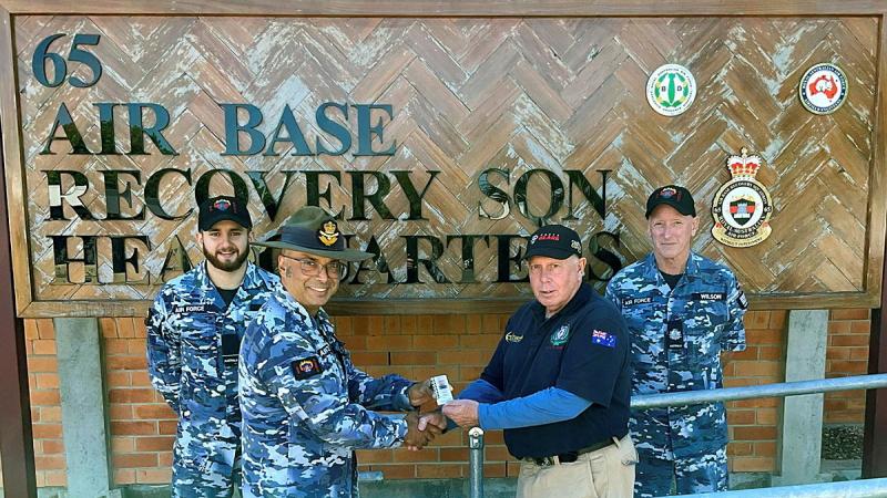 Military personnel give a small item to a civilian in front of a 65 Air base Recovery SQN Headquarters sign.