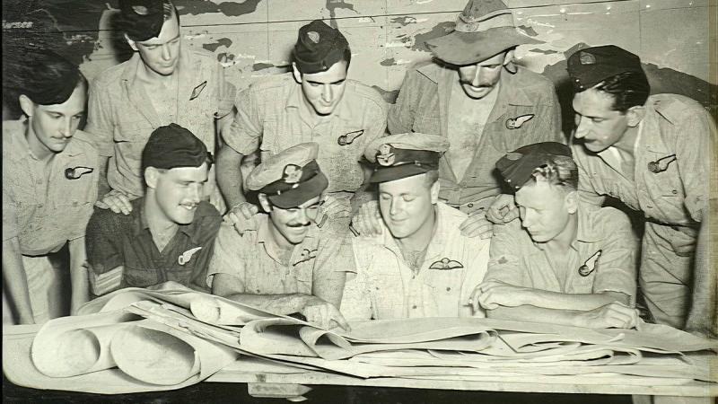 A black-and-white historical image of a group of men gathered around a table looking at maps.