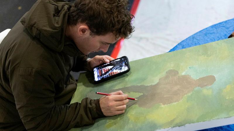 A man holds a phone in one hand while drawing on a painted canvas with a graphite pencil in the other.