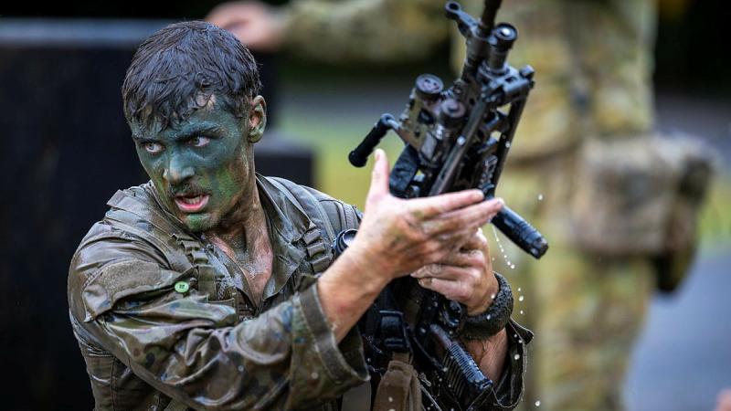 A soaked man in camouflage uniform and face paint holds a rifle as water splashes from it.