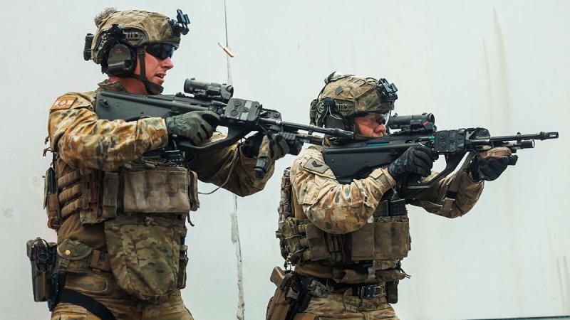 Two men in uniform move alongside a wall with rifles raised, a bullet casing in the air next to one of them.