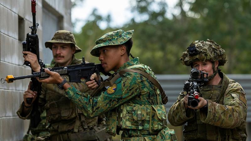 Three people in camouflage uniform hold machine guns, two of them aiming the guns, as they walk next to a cinderblock building.