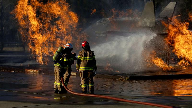 Three people in firefighting uniforms directing the spray from a fire hose at an aircraft in flames.