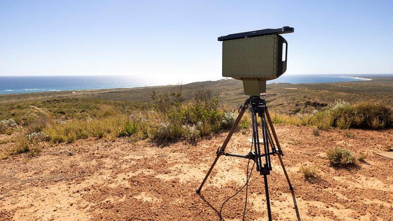 A rectangular device on a tripod standing in red soil, with short scrub nearby and the sea in the distance.
