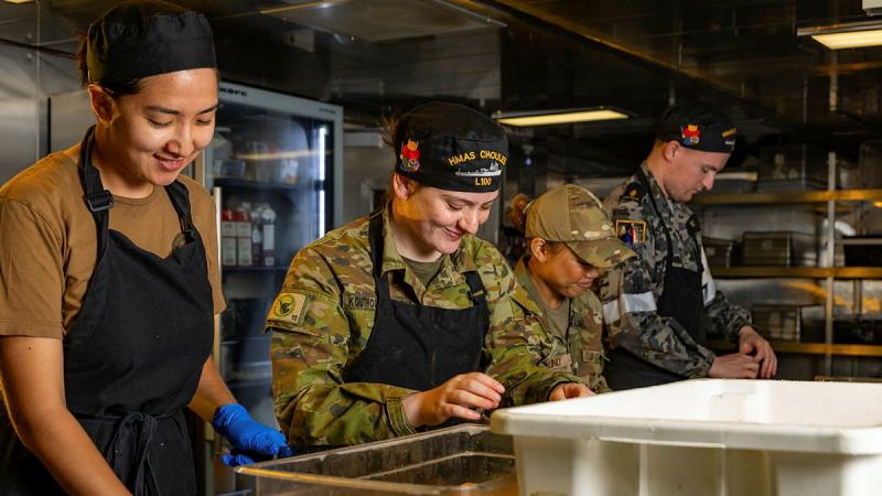 Military personnel prepare food in an industrial kitchen.