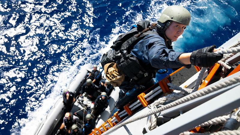 A woman wearing a helmet and gloves climbs up the side of a ship by a rope ladder, with a small boat in the water carrying people directly beneath her.