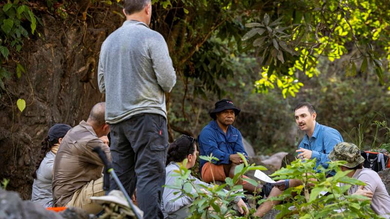 A group of people sit and stand amongst vegetation facing a seated man who is speaking.
