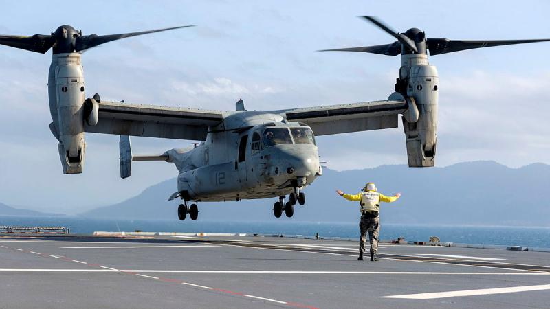A defence member in a bright yellow uniform signal for an aircraft to land on the deck of a ship.