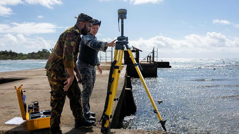 Two people in uniform examine a device on a tripod on a jetty.