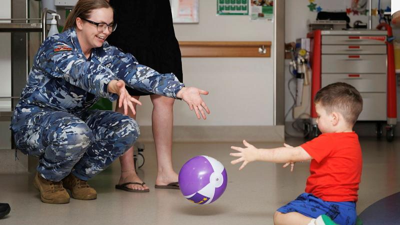 A military woman throws a purple ball to a young child kneeling on the floor.
