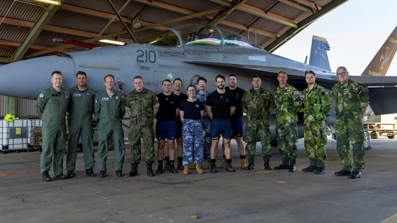 A group of aviators from Sweden, Finland and Australia stand in front of a jet aircraft and smile at the camera.