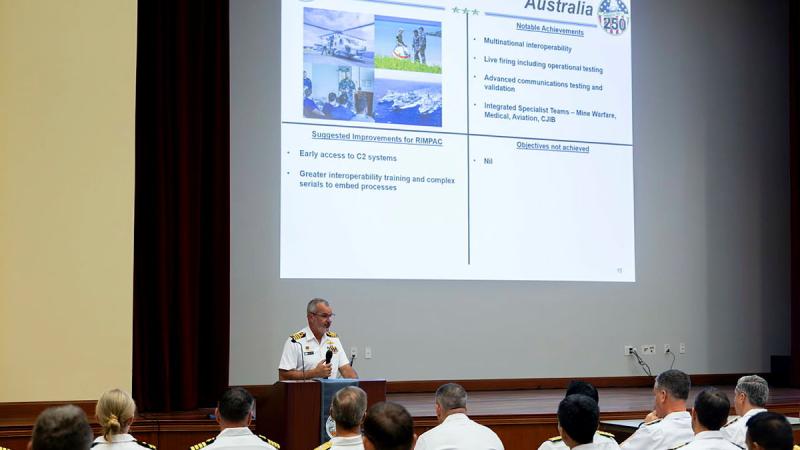 A man in a white naval uniform speaks at a lectern while an audience watches a presentation on a screen behind him.