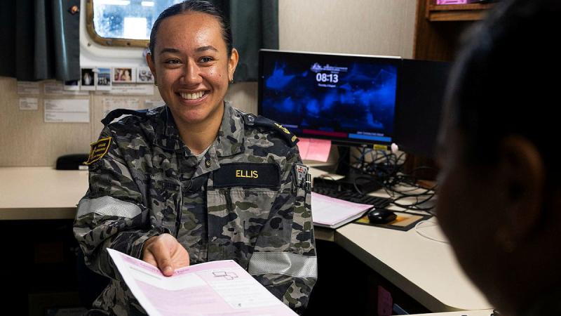 A woman in uniform sits in an office and takes a form handed to her by another person.