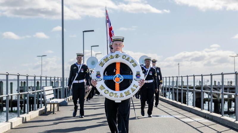 A person in Navy uniform carries a lifebuoy along a jetty with a group of uniformed people behind.