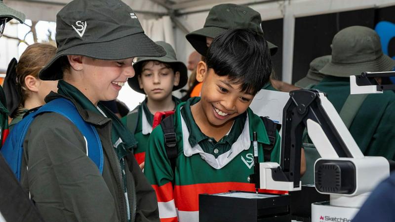 Several young kids inspect a 3D printing machine.