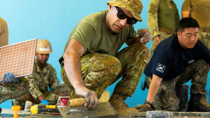 A man in Army uniform crouches while using a tile trowel, with others working around him.