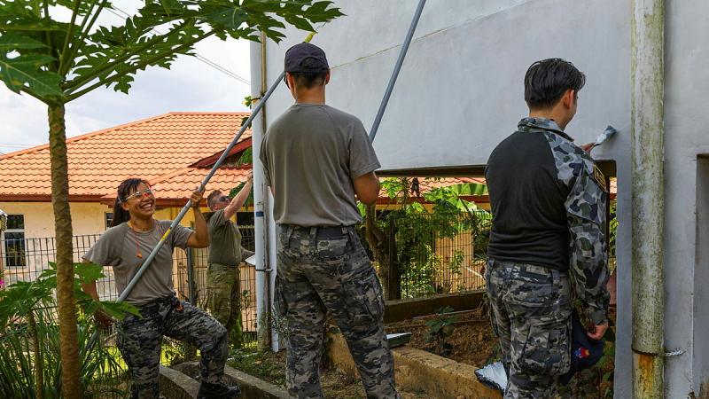 Three people in Navy uniform paint a building wall with brushes and rollers while a person in Army uniform paints another wall.