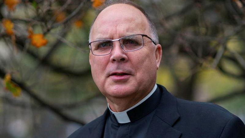 A man in a black suit and clerical collar stands outside.